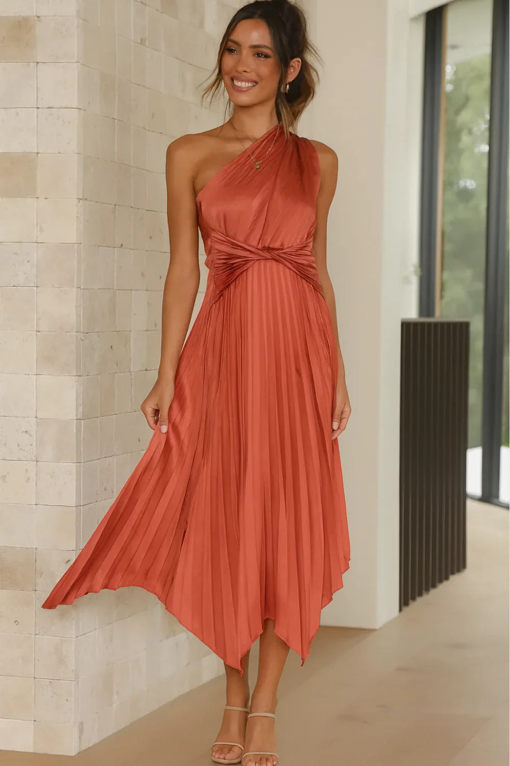 Woman wearing Kouvr's Audrine one-shoulder pleated midi dress in copper, showcasing the twisted waist and flowing asymmetrical hem
