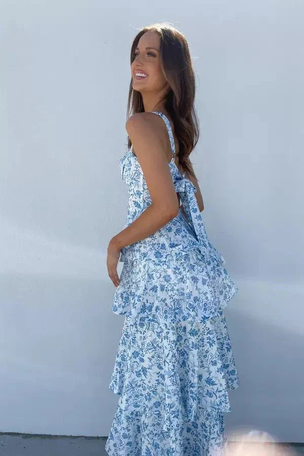 Model wearing the Kouvr Denise blue and white floral tiered midi dress with spaghetti straps, standing against a white wall.