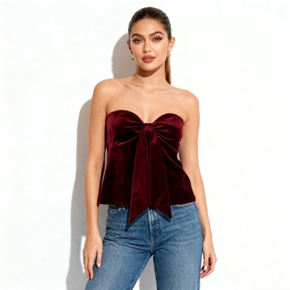 Model wearing the Sophia burgundy velvet bow strapless top from Kouvr Fashion, paired with high-waisted blue jeans against a white background.