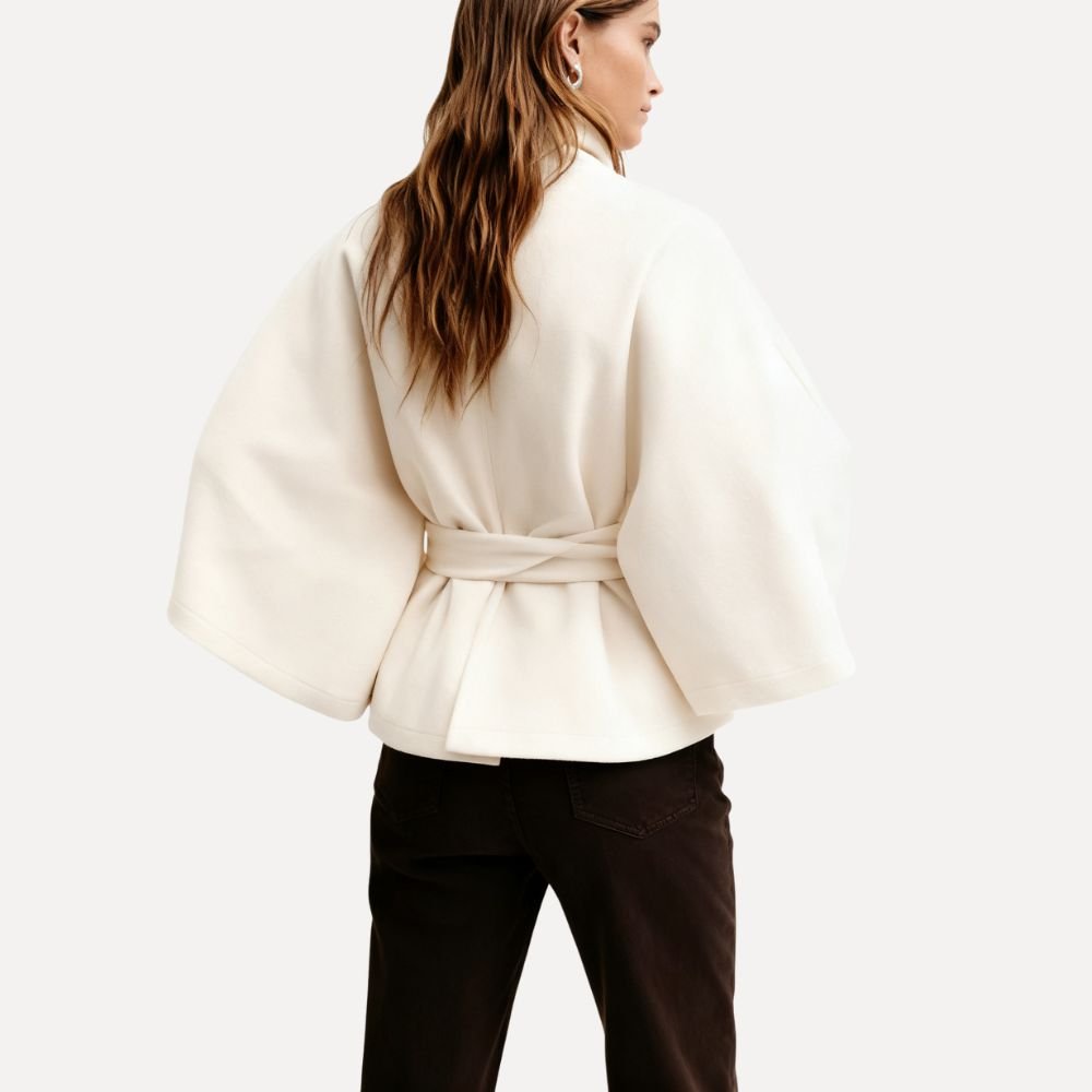 Model wearing the Kouvr Jannah Wool Cape Jacket in White, featuring a high funnel neck and tie-waist belt." For the back view