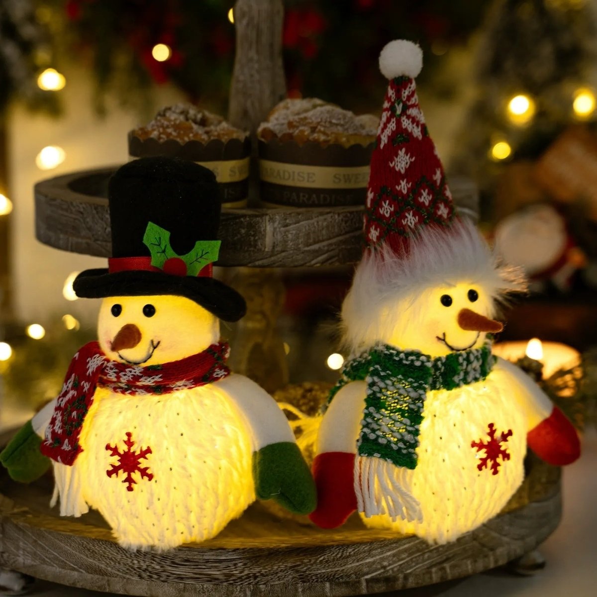 A pair of Kouvr's light-up plush snowman figurines glowing warmly on a rustic wooden tiered tray, surrounded by festive Christmas lights and muffins.