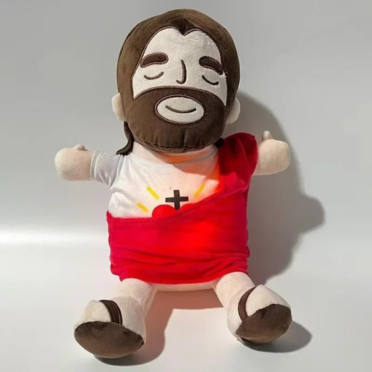 Studio product shot of the Kouvr interactive Jesus plush soother with a red sash and glowing Sacred Heart, sitting on a white background.