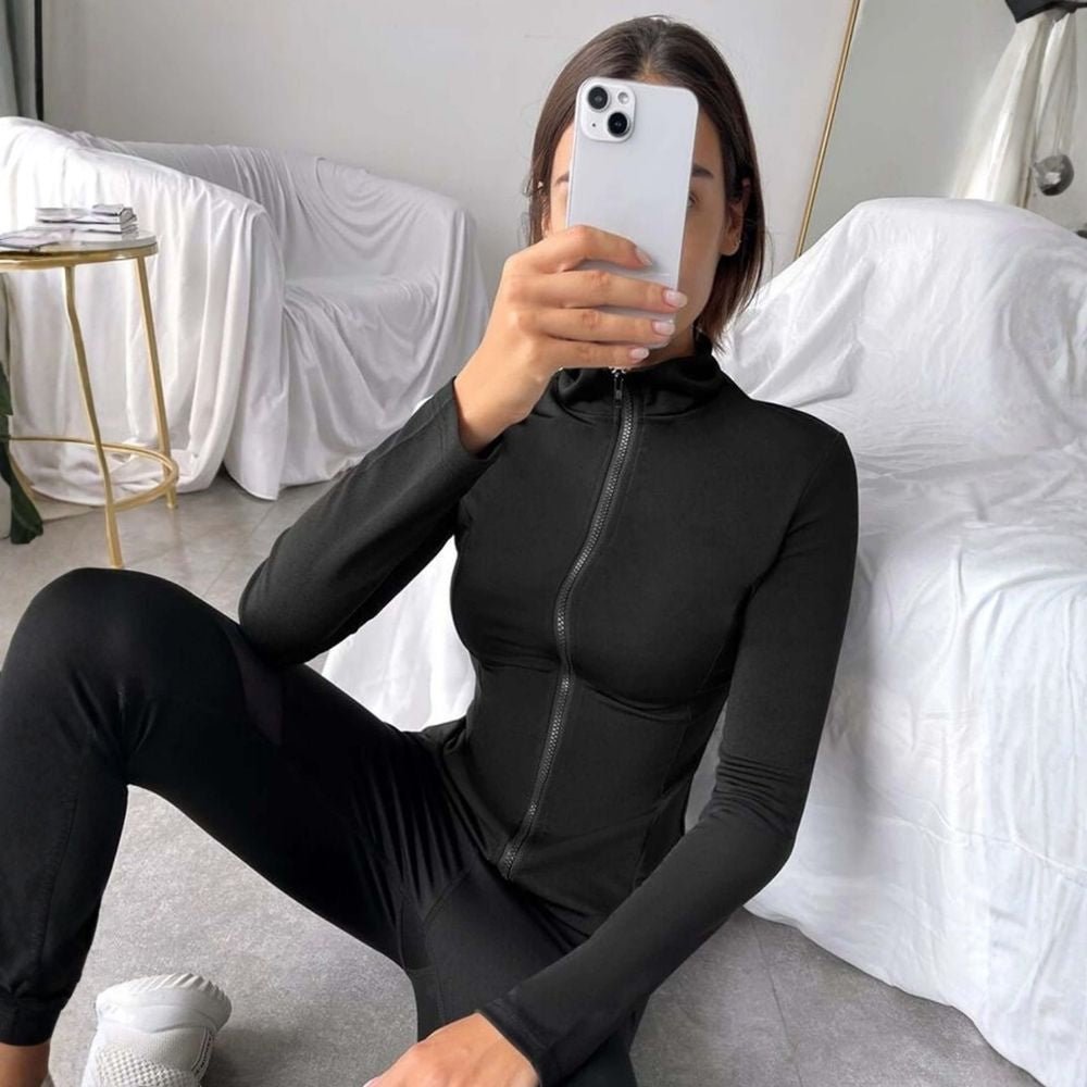 Woman wearing the black Kouvr Sculpting Athletic Jacket in a modern fitness studio.