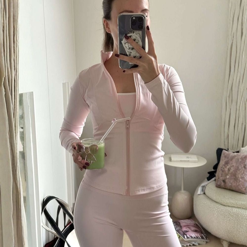 Woman wearing the Kouvr Sculpting Zip Jacket in Petal Pink against a white wall.