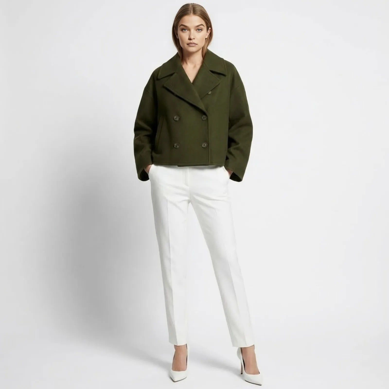 A female model wearing the Kouvr dark green cropped wool peacoat, styled with white tailored trousers and white heels, seen from the front.