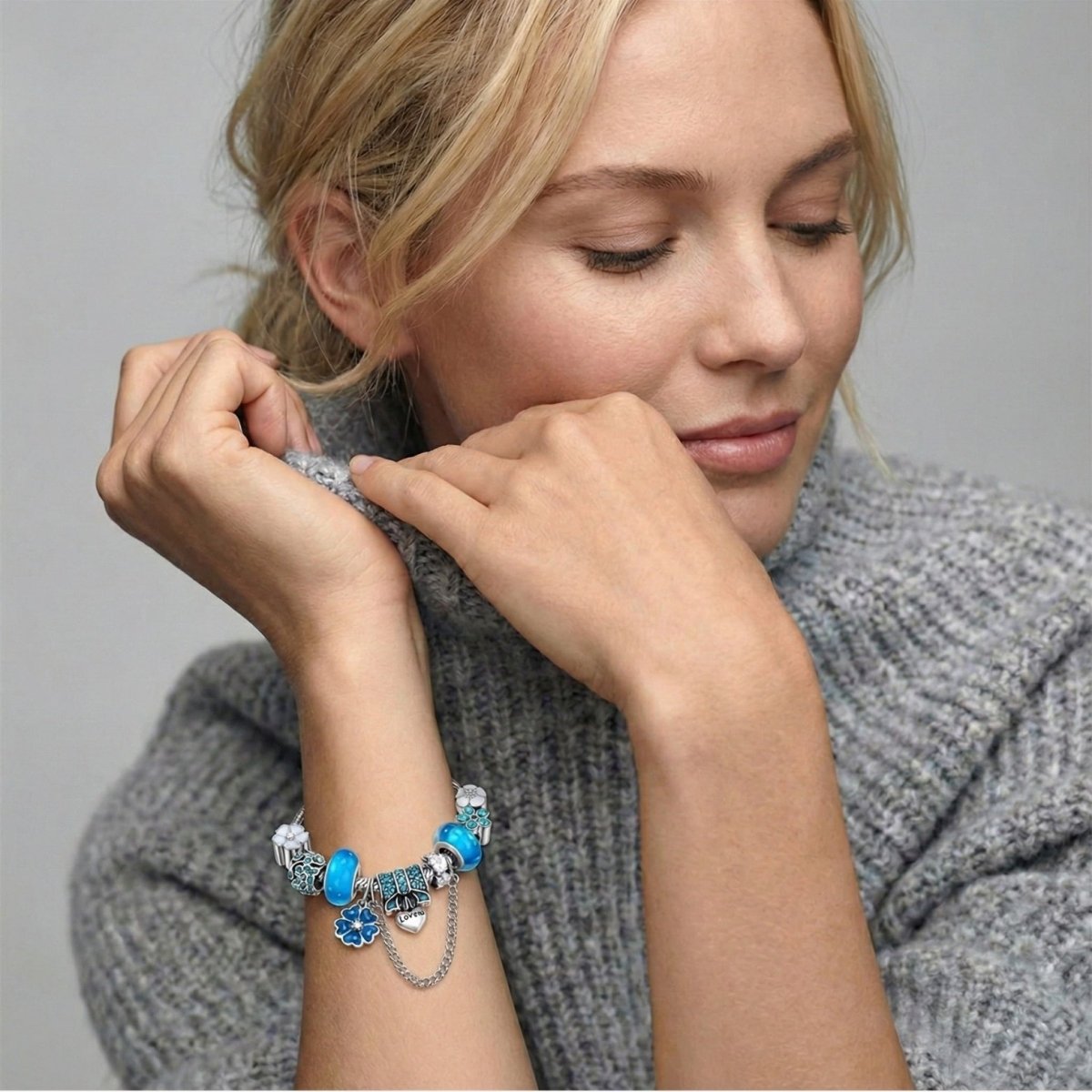Blonde woman wearing a grey knit turtleneck sweater, resting her chin on her hands, showcasing a silver charm bracelet with blue beads and a "Love" heart charm.