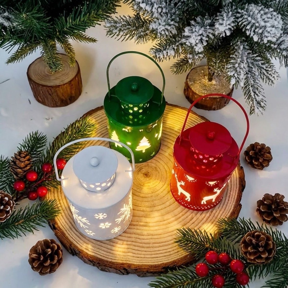 Top-down view of a set of three Christmas lanterns (red, green, white) arranged on a natural wood slice, surrounded by frosted mini trees and pinecones.