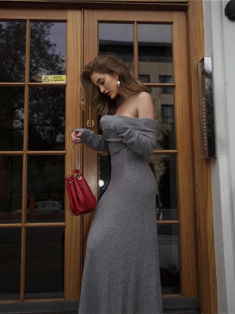 A woman in the Kouvr Ingrid grey off-the-shoulder sweater dress looking down at her red handbag in front of a wooden and glass door.
