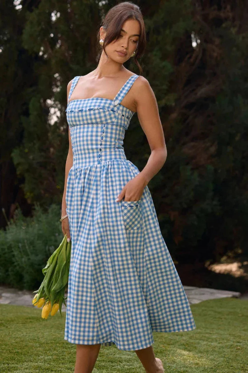 Model wearing the Kouvr Apolline blue gingham midi dress with a structured corset bodice and square neckline, standing in a garden
