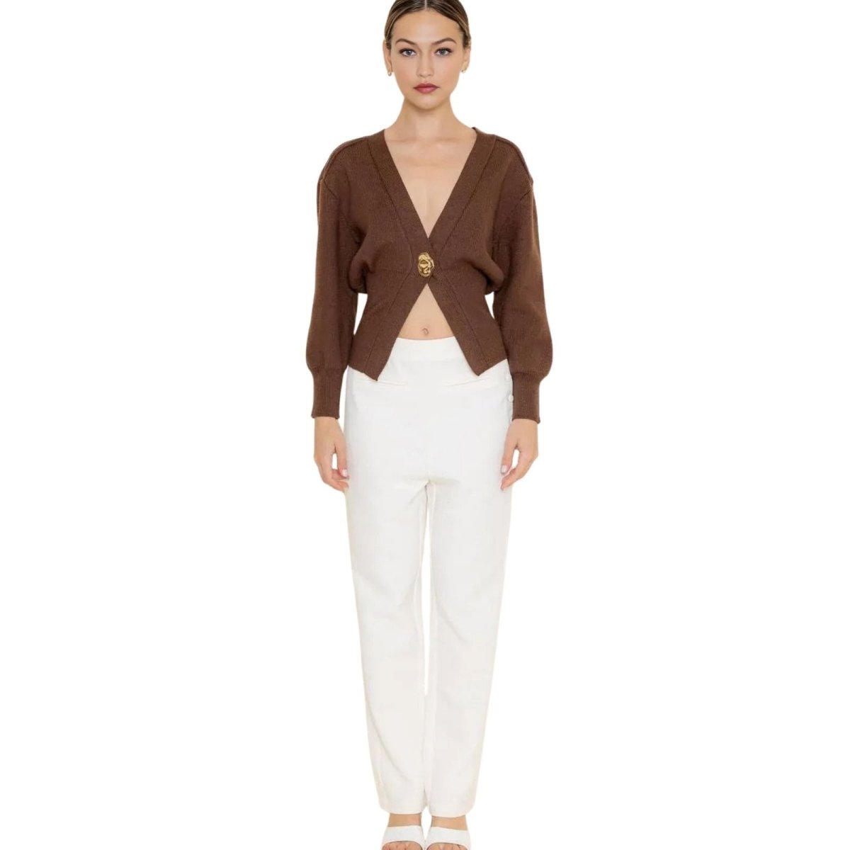 Woman wearing a brown kouvr cardigan over a white top with white pants on a white background