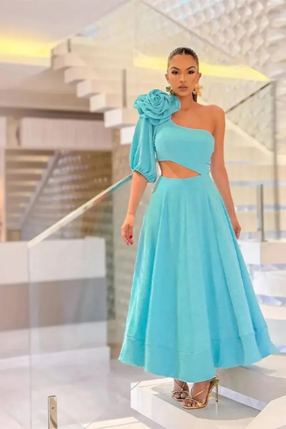 Model standing on a staircase wearing the Kouvr light blue one-shoulder dress with a large flower detail and side cutout