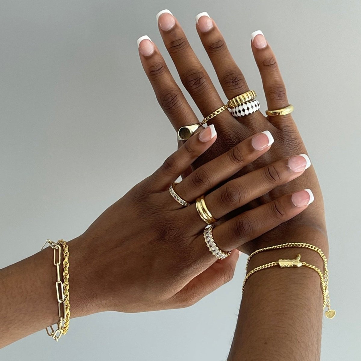 Two hands wearing multiple gold kouvr rings and bracelets on a neutral background
