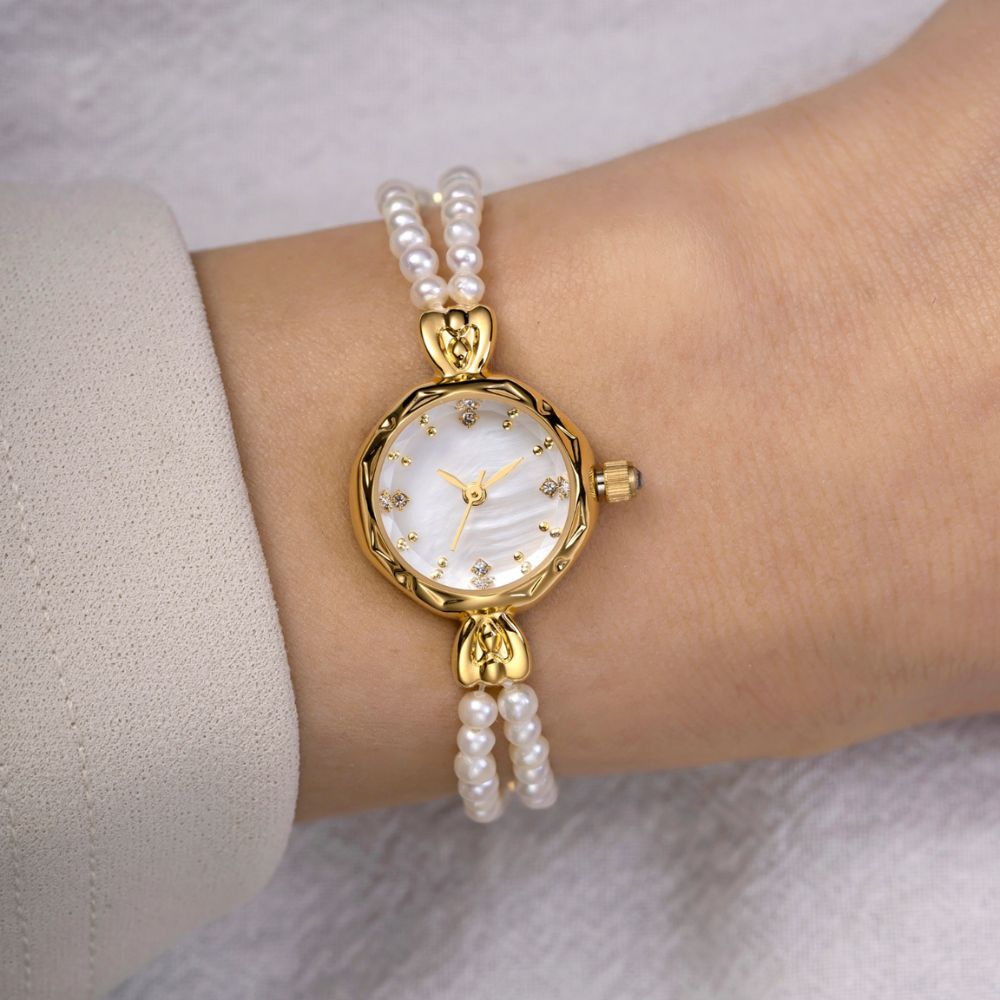 Gold kouvr watch with pearl bracelet on a wrist against a neutral background