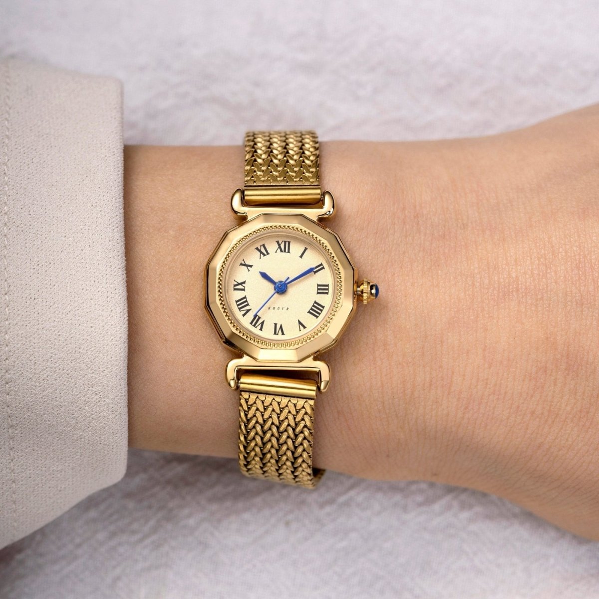 Gold Kouvr wristwatch with a white face and blue hands on a wrist against a neutral background