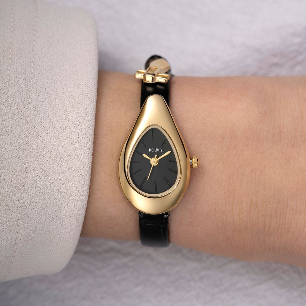 Gold watch with black kouvr strap on a wrist against a neutral background