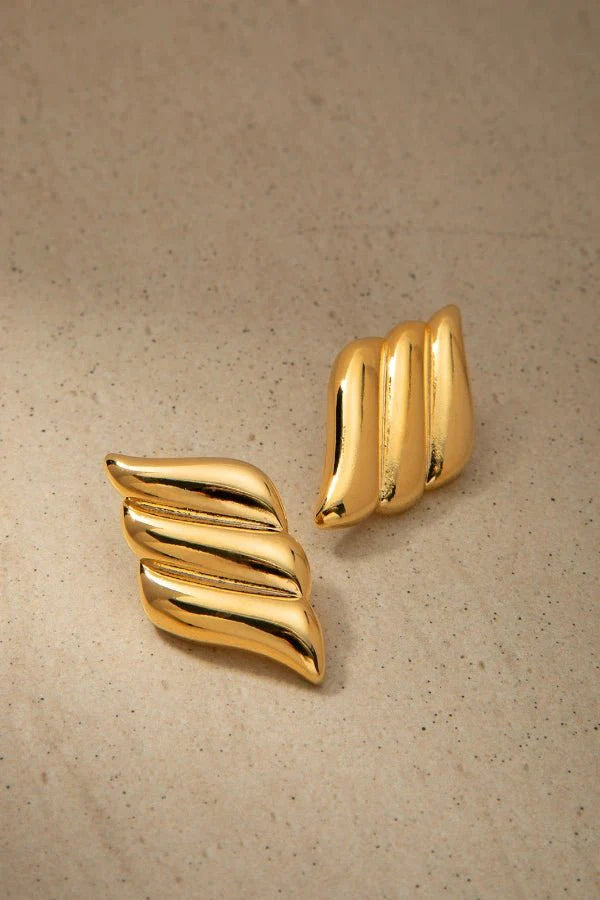 A product shot of a pair of Kouvr Fashion gold wavy statement earrings laid on a textured neutral surface.