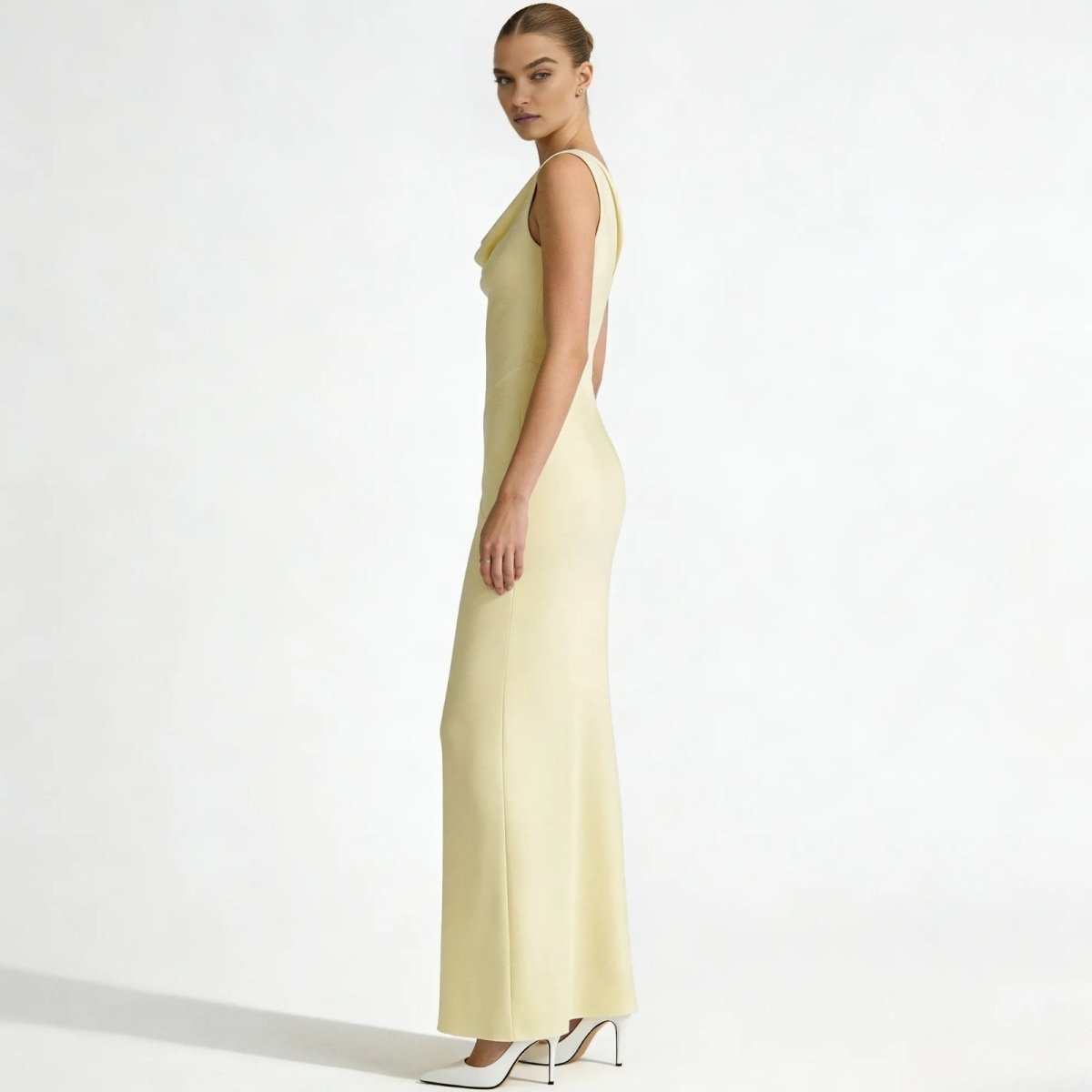 Side profile of a model in the Kouvr Fashion Vickie Gown, showcasing the elegant column silhouette and draped cowl neckline of the pale ivory dress.
