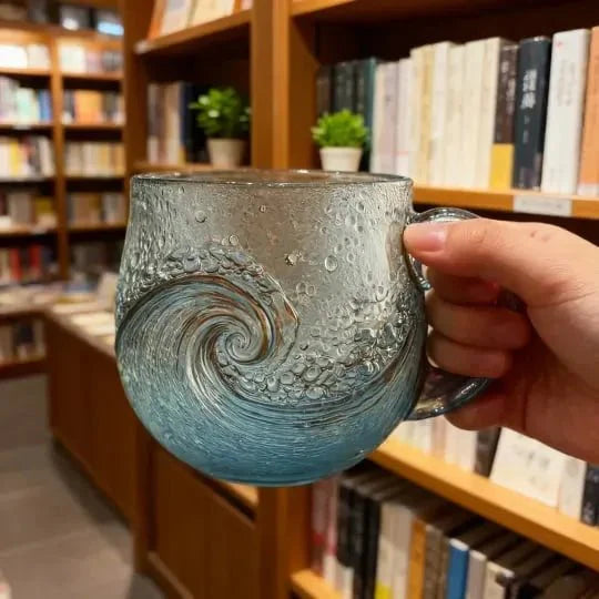 A hand holding the textured Kouvr ocean wave mug, showing the detail of the blue swirl design and bubbly glass.