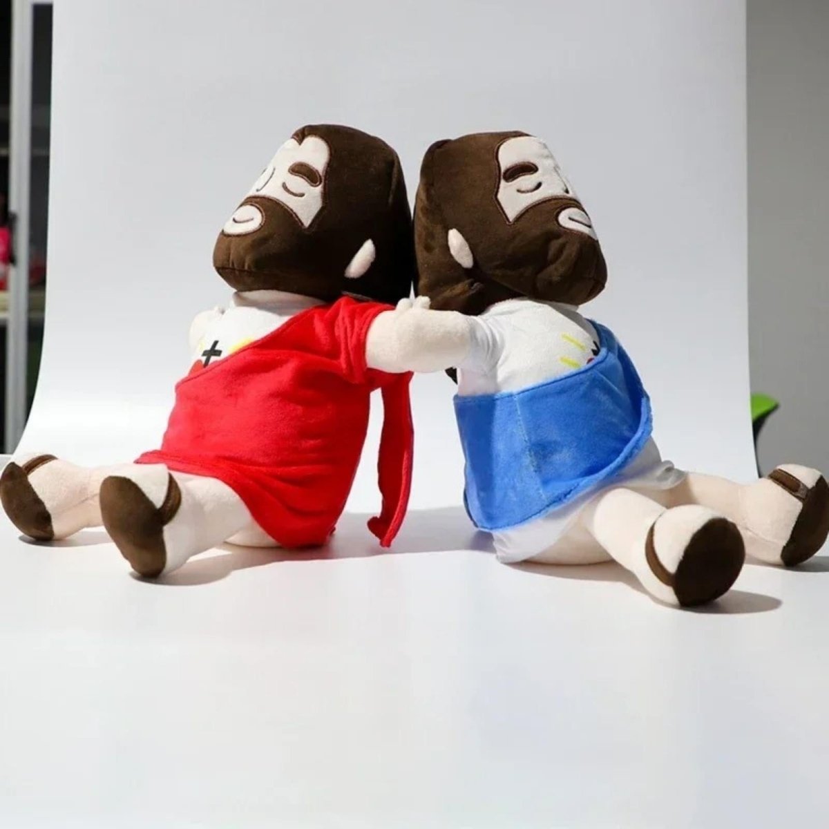 The back view of two Kouvr Jesus plush dolls sitting back-to-back with their arms interlinked, symbolizing friendship and unity.