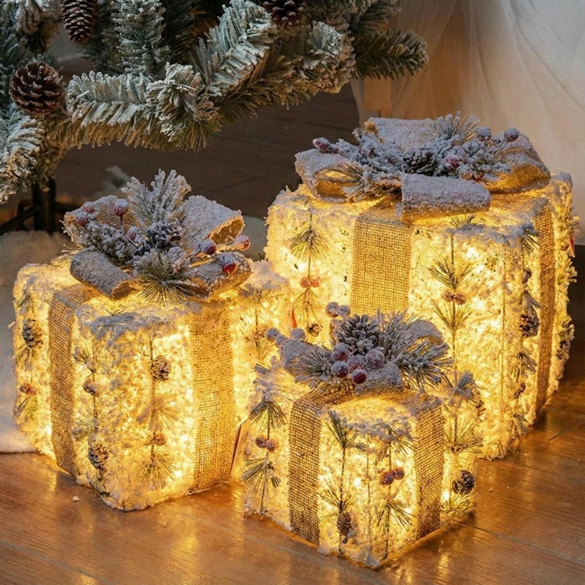 Close-up of three pre-lit Christmas gift boxes decorated with burlap, flocked pine, and berries, nestled under Christmas tree branches.