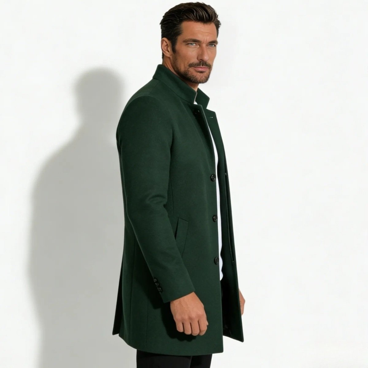 Side profile of a man in a slim fit dark green wool topcoat with a stand collar, showing the coat's silhouette and length.
 