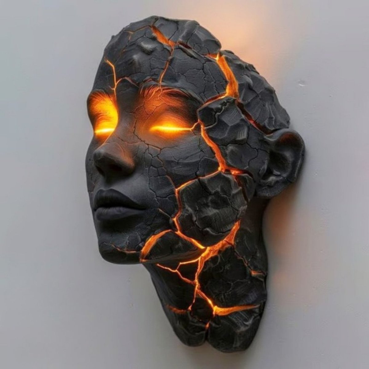 Angled view of the Sculptural Wall Sconce by Kouvr, showing the deep texture of the black cracked lava stone face and the warm orange glow from the eyes and fissures.