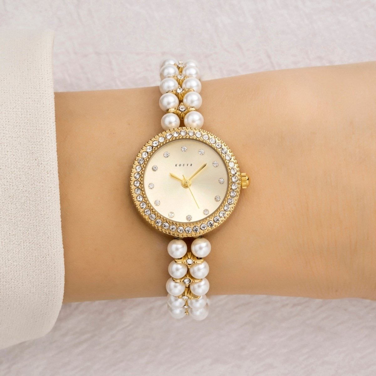 Gold Kouvr watch with pearl bracelet on a wrist against a light background