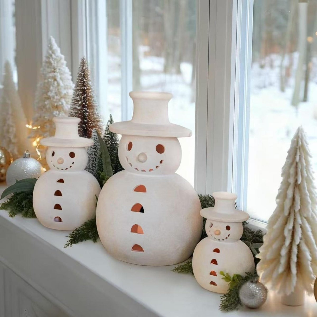 A family of three white ceramic snowman luminaries styled on a bright windowsill, surrounded by miniature Christmas trees and winter greenery, with a snowy landscape visible outside.