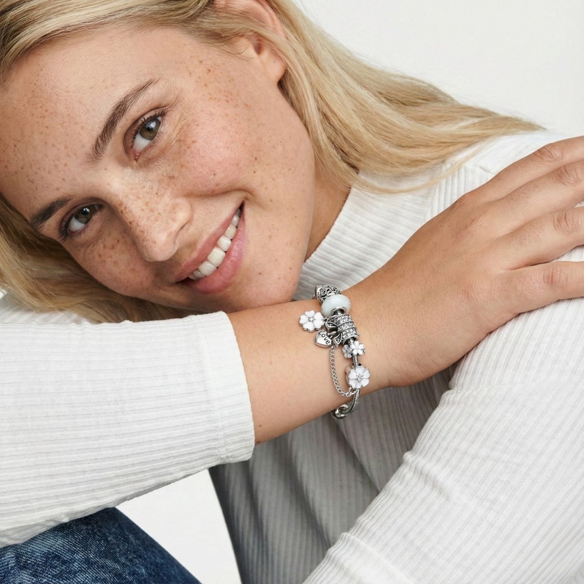 Smiling blonde woman with freckles wearing a white ribbed top, displaying a silver charm bracelet with white flower charms and glass beads on her wrist.
